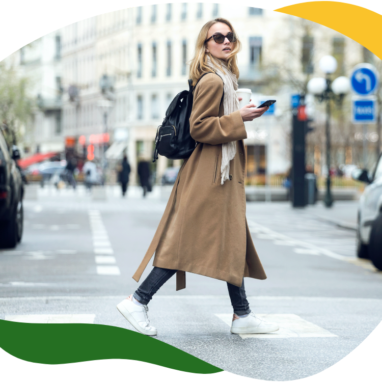 A modern woman with big sunglasses and a beige long coat crosses the street holding her mobile phone in her right hand