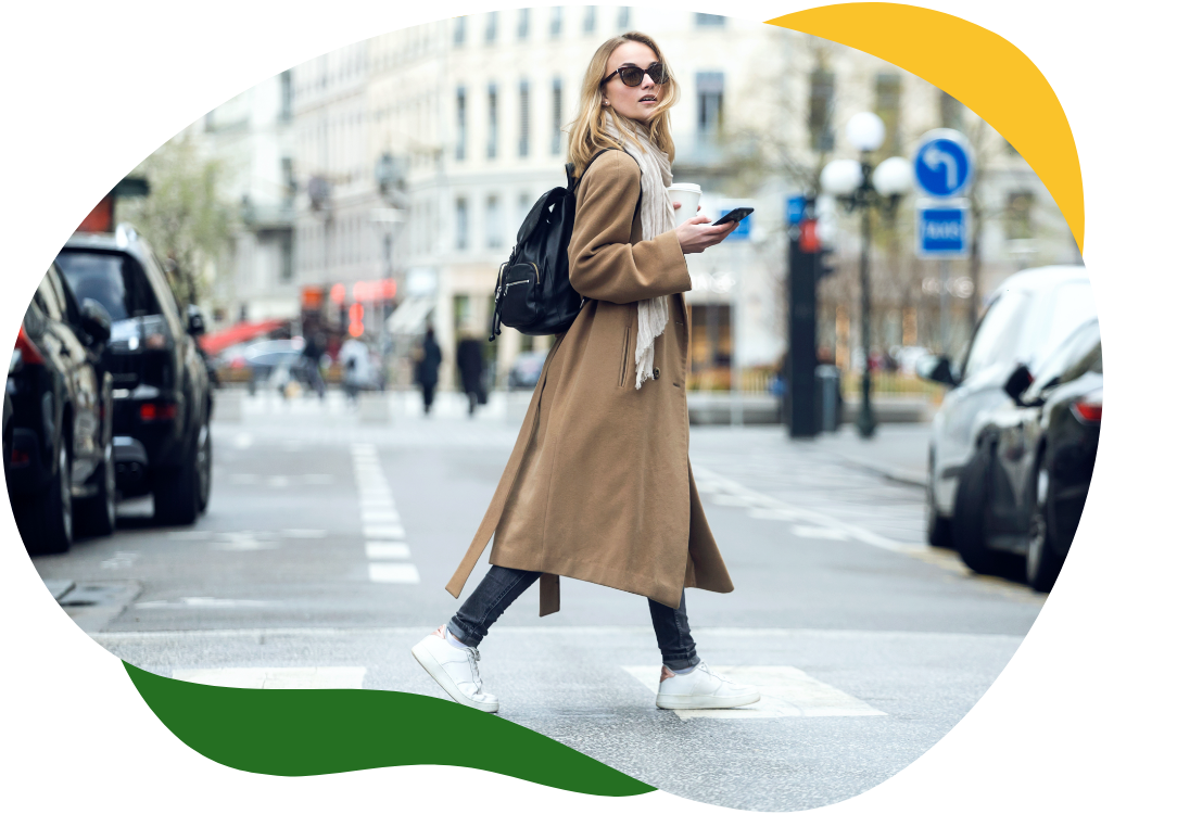 A modern woman with big sunglasses and a beige long coat crosses the street holding her mobile phone in her right hand